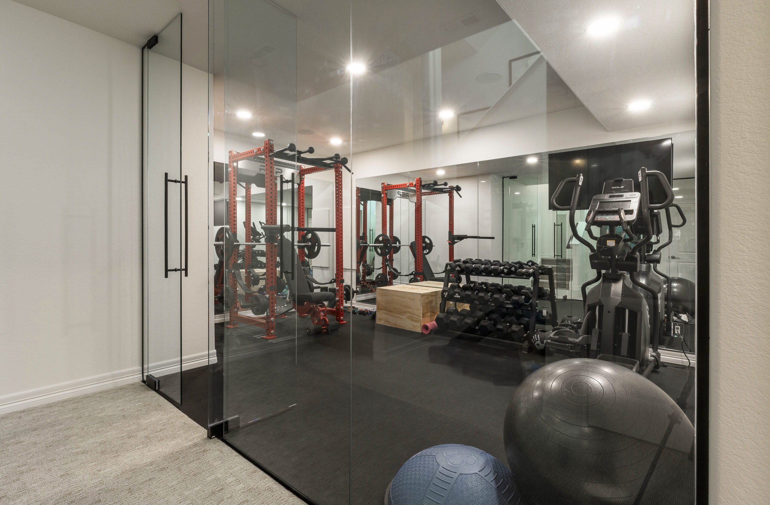 Glass-enclosed gym area with various exercise equipment, including a treadmill, weights, and a power rack, in a renovated basement.